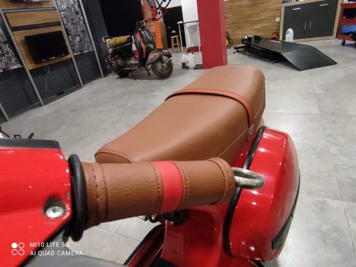 Asiento de moto roja con tapizado marrón y detalles en rojo. Fondo de un taller.
