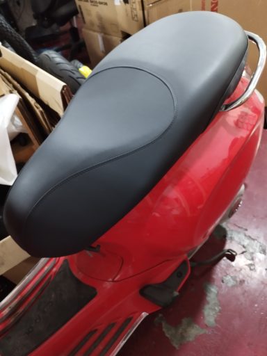 Asiento negro de una scooter roja, sobre fondo desordenado de objetos.