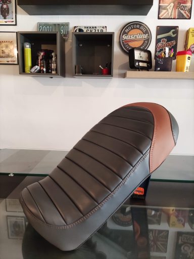 Asiento de motocicleta con diseño negro y marrón, sobre una superficie plateada.