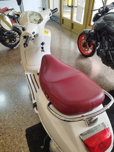 Escúter blanco con asiento rojo, situado en un concesionario de motos.
