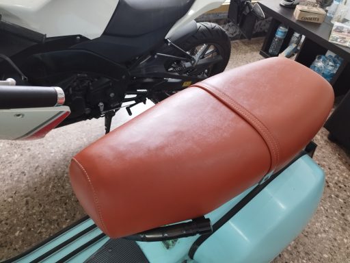 Asiento de moto de color marrón, con superficie lisa, sobre un fondo de muebles y un vehículo.