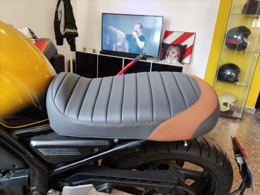 Asiento de motocicleta con diseño acolchado en tonos gris y marrón. Fondo con televisión.